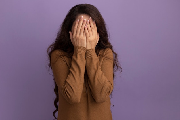 Young beautiful girl wearing brown turtleneck sweater covered face with hands isolated on purple wall