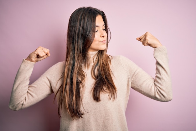 Free photo young beautiful girl wearing casual turtleneck sweater standing over isolated pink background showing arms muscles smiling proud fitness concept