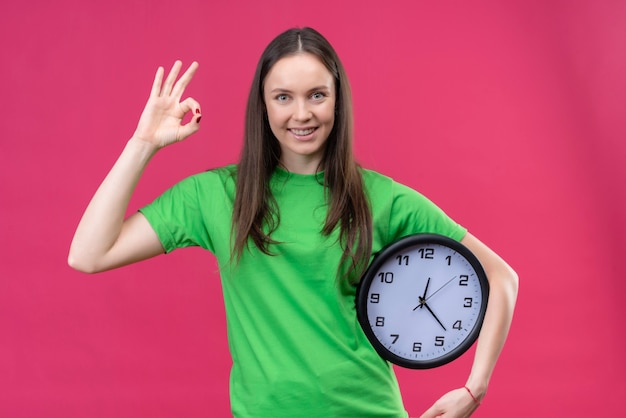 Giovane bella ragazza che indossa la maglietta verde che tiene l'orologio che sorride allegramente facendo segno giusto che sta sopra fondo rosa isolato