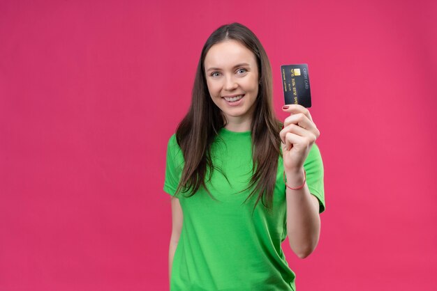 Young beautiful girl wearing green t-shirt holding credit card smiling cheerfully happy and positive standing over isolated pink background