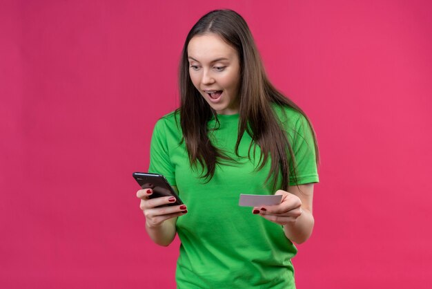 Молодая красивая девушка в зеленой футболке держит смартфон, глядя на экран, изумлен и удивлен, стоя на изолированном розовом фоне