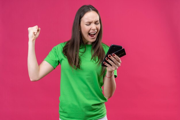 Young beautiful girl wearing green t-shirt holding smartphone looking at screen exited and happy raising clenched fist smiling cheerfully rejoicing her success standing over isolated pink bac