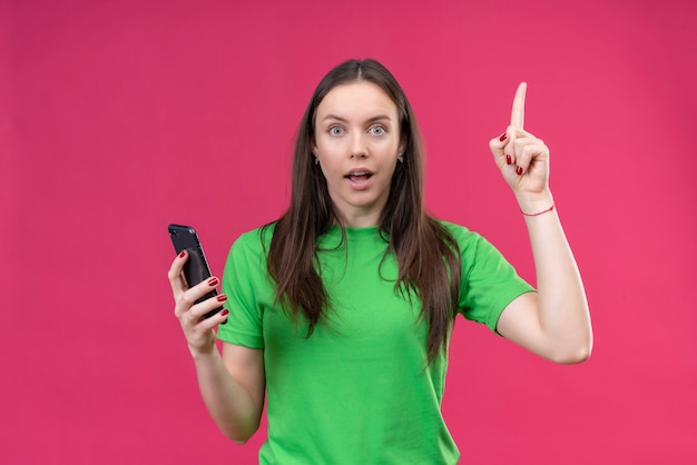 孤立したピンクの背景の上に立っている新しいアイデアコンセプトを持つスマートフォンの人差し指を保持している緑のtシャツを着ている美しい少女