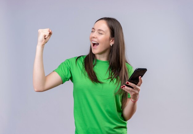 Молодая красивая девушка в зеленой футболке держит смартфон, поднимая сжатый кулак, счастлива и выходит, улыбаясь, радуясь своему успеху, стоя на изолированном белом фоне