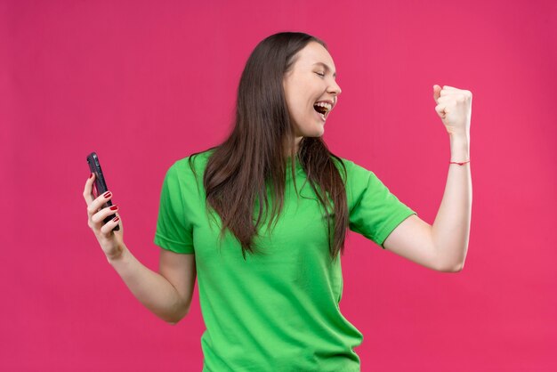 Young beautiful girl wearing green t-shirt holding smartphone raising fist crazy happy rejoicing her success standing over isolated pink background