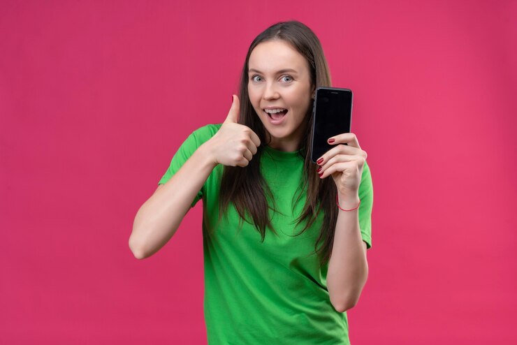 Free Photo young beautiful girl wearing green t-shirt holding smartphone smiling cheerfully showing thumbs up standing over isolated pink background