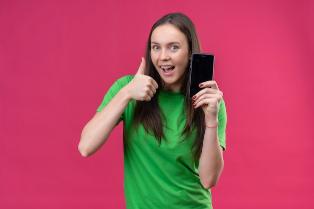 Young beautiful girl wearing green t-shirt holding smartphone smiling cheerfully showing thumbs up standing over isolated pink background