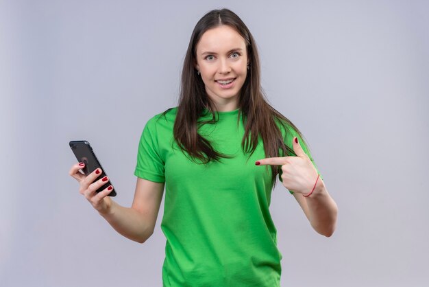 Young beautiful girl wearing green t-shirt holding smartphone smiling pointing with finger to it standing over isolated white background