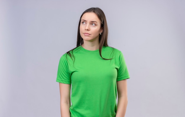 Молодая красивая девушка в зеленой футболке недовольно смотрит в сторону, стоя на изолированном белом фоне