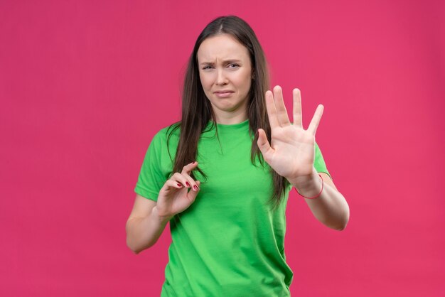 Young beautiful girl wearing green t-shirt making defense gesture with open hands with disgusted expression on face holding hands up telling do not come closer standing over isolated pink ba