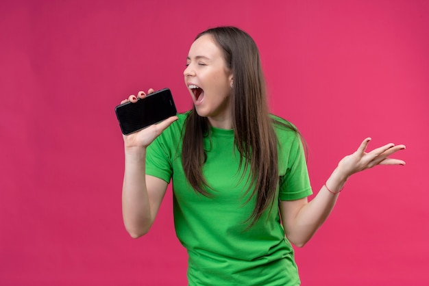 격리 된 분홍색 배경 위에 서있는 마이크로 사용하는 그녀의 스마트 폰에 외치는 녹색 티셔츠를 입고 젊은 아름다운 소녀