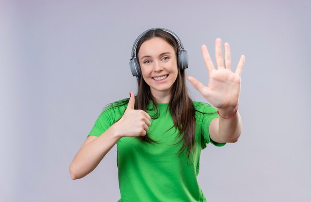 Молодая красивая девушка в зеленой футболке весело улыбается, показывая пальцами номер пять и поднимает вверх большие пальцы руки, весело улыбаясь, стоя на изолированном белом фоне