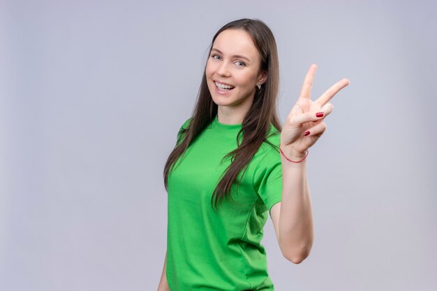緑のtシャツを着て美しい少女元気に笑みを浮かべて表示し、分離された白い背景の上に立っている指番号2または勝利のサインで上向き