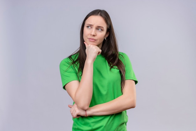 Молодая красивая девушка в зеленой футболке стоит с рукой на подбородке, глядя в сторону с задумчивым выражением лица, стоящего на изолированном белом фоне