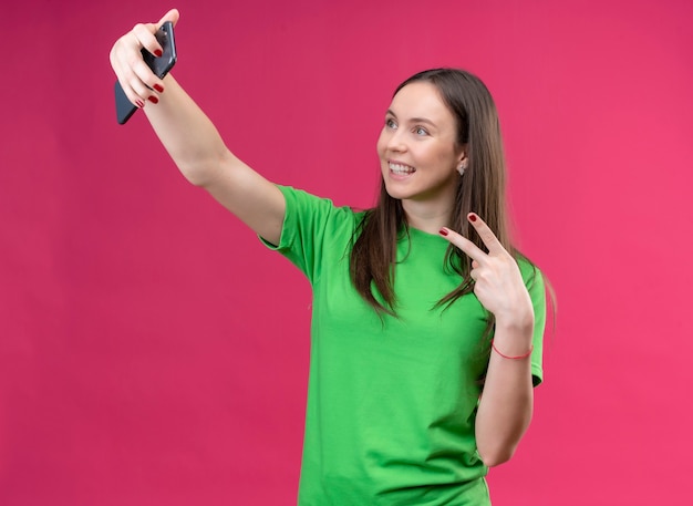 孤立したピンクの背景の上に元気に立っている笑顔の勝利のサインを示すスマートフォンを使用してselfieを取って緑のtシャツを着ている美しい少女