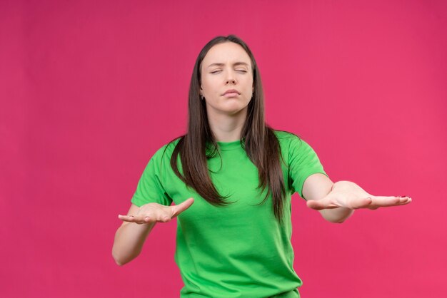 Young beautiful girl wearing green t-shirt walking with closed eyes holding out her hands ti find road standing over isolated pink background