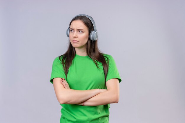 Young beautiful girl wearing green t-shirt with headphones standing with arms crossed looking aside displeased over isolated white background