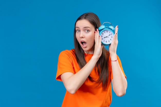 Young beautiful girl wearing orange t-shirt holding alarm clock looking amazed and surprised standing over isolated blue background