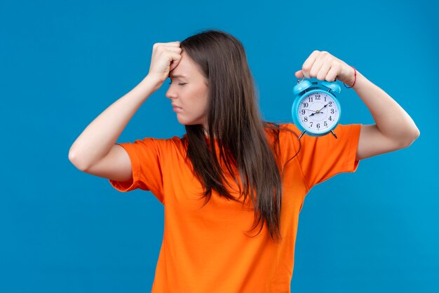 Young beautiful girl wearing orange t-shirt holding alarm clock touching her head for mistake bad memory concept standing over  isolated blue background