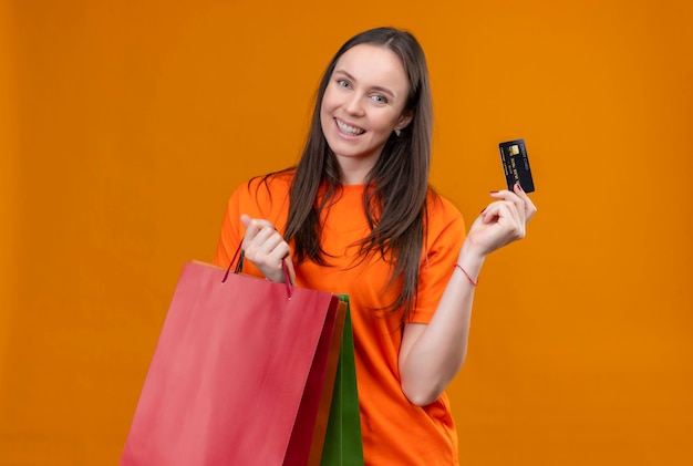 紙袋と孤立したオレンジ色の背景の上に元気に立っている笑顔のクレジットカードを保持しているオレンジ色のtシャツを着ている美しい少女