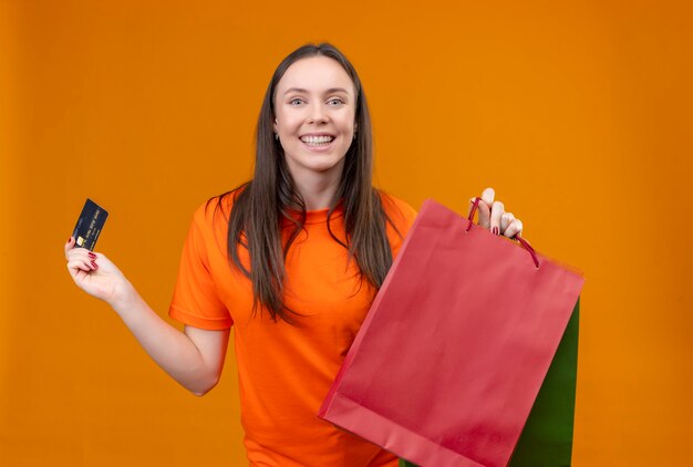 Молодая красивая девушка в оранжевой футболке держит бумажный пакет и кредитную карту, весело улыбаясь, стоя на изолированном оранжевом фоне