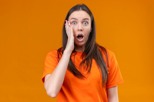 Young beautiful girl wearing orange t-shirt looking amazed and surprised touching her face with hand standing over isolated orange background