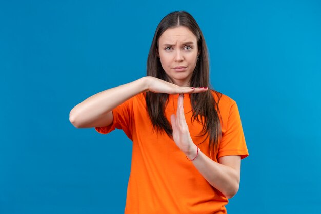 Молодая красивая девушка в оранжевой футболке недовольно жестикулирует руками, делая жест тайм-аута, стоя на изолированном синем фоне