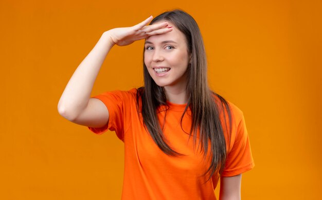 孤立したオレンジ色の背景の上に立っている笑顔の誰かを見て頭の上の手で遠く離れているオレンジ色のtシャツを着ている美しい少女