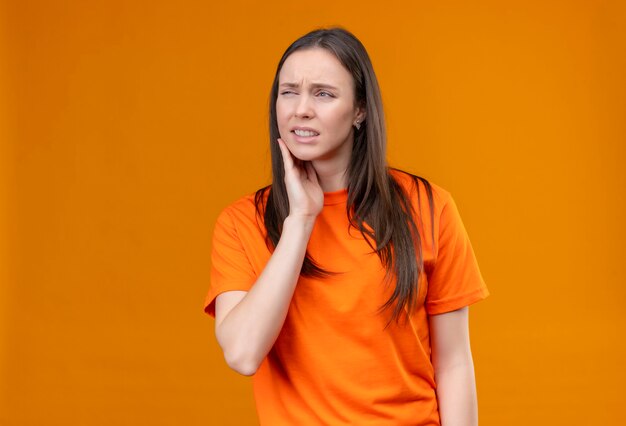 Young beautiful girl wearing orange t-shirt looking unwell touching her cheek feeling toothache standing over isolated orange background