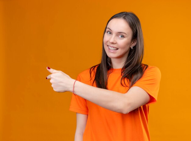 Young beautiful girl wearing orange t-shirt pointing to the side smiling cheerfully standing over isolated orange background