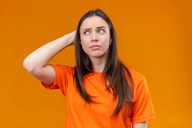 Молодая красивая девушка в оранжевой футболке почесывает голову, глядя в сторону с задумчивым подозрительным выражением лица, стоящего на изолированном оранжевом фоне