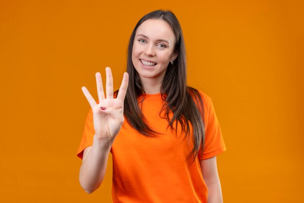オレンジ色のtシャツを着て、孤立したオレンジ色の背景の上に立っている指番号4を上向きに身に着けている美しい少女