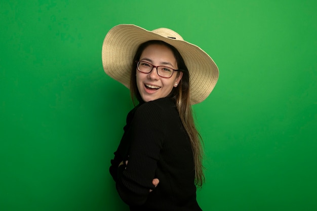 Giovane bella ragazza che indossa il cappello estivo in un dolcevita nero e occhiali sorridendo felice e positivo allegramente