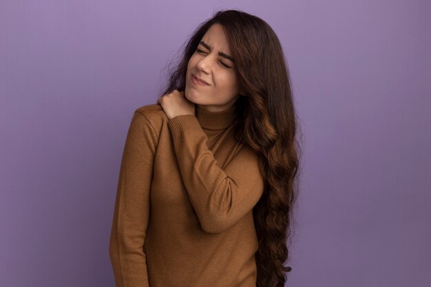 Young beautiful girl with closed eyes wearing brown turtleneck sweater grabbed aching shoulder isolated on purple wall