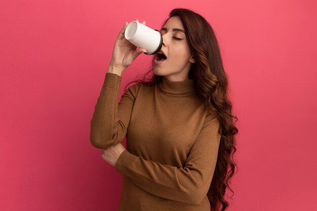 Giovane bella ragazza con gli occhi chiusi che indossa il maglione a collo alto beve caffè isolato sulla parete rosa