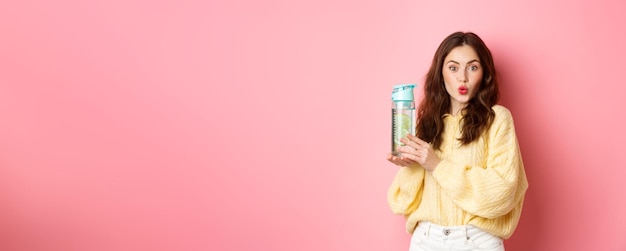 곱슬머리 입술을 가진 젊은 미녀와 분홍색 배경에 서있는 레몬 물병 음료수를 보여주는 흥분한 모습