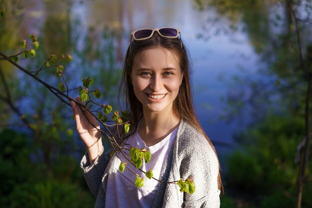 A young beautiful girl with sunglasses is walking near the lake, holding a tree branch in her hands