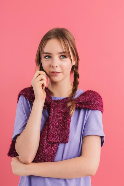 Young beautiful girl with two braids in lilac t shirt and sweater on shoulders talking on cellphone while happily looking aside over pink background