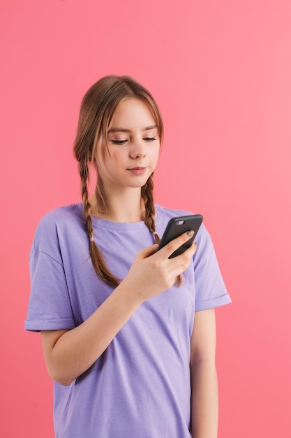 Молодая красивая девушка с двумя косами в сиреневой футболке задумчиво пользуется мобильным телефоном, проводя время на розовом фоне