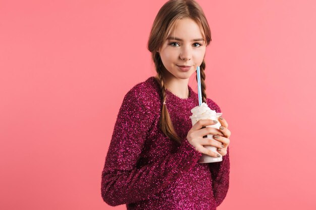 Young beautiful girl with two braids in sweater holding milkshake in hands while happily looking in camera over pink background