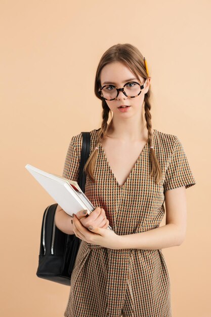 Young beautiful girl with two braids in tweed jumpsuit and eyeglasses with black backpack on shoulder and pencil behind ear holding book in hand while dreamily looking in camera over beige background