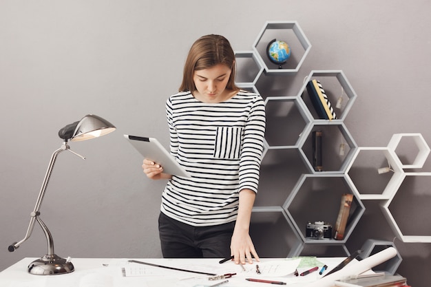 Young beautiful good-looking dark-haired girl in striped shirt standing near table in comfy workplace at home, holding digital tablet in hands, looking at papers with concentrated expression, trying t