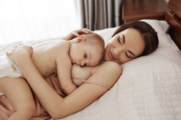 Молодая красивая счастливая грудное вскармливание мамы обнимая ее младенца лежа на кровати дома.