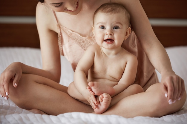Бесплатное фото Молодая красивая счастливая мама в пижамы и ее новорожденного ребенка, сидя на кровати в утро, улыбаясь, играть вместе.