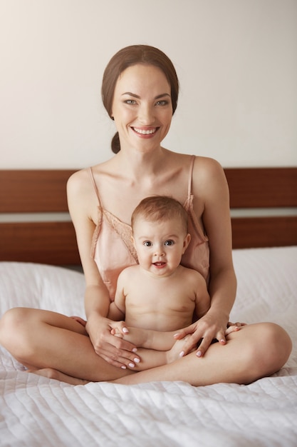 Young beautiful happy mom in sleepwear and her newborn baby sitting on bed in morning smiling playing together.