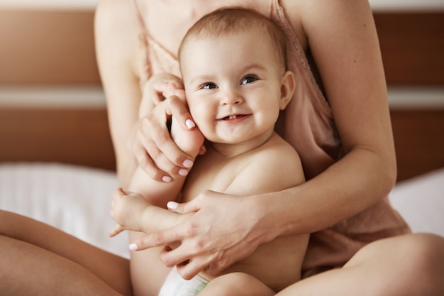 Young beautiful happy mom in sleepwear and her newborn baby sitting on bed smiling playing together at home.