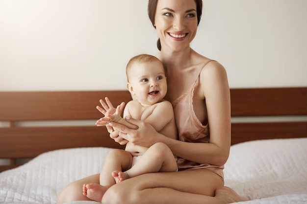 Young beautiful happy mother in sleepwear and her newborn baby sitting on bed in morning smiling playing together.