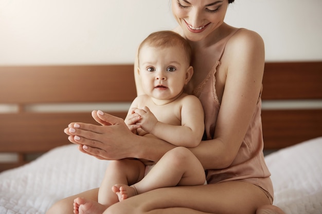 Молодая красивая счастливая мать в пижамы и ее новорожденного ребенка, сидя на кровати в утренней улыбкой, играть вместе.
