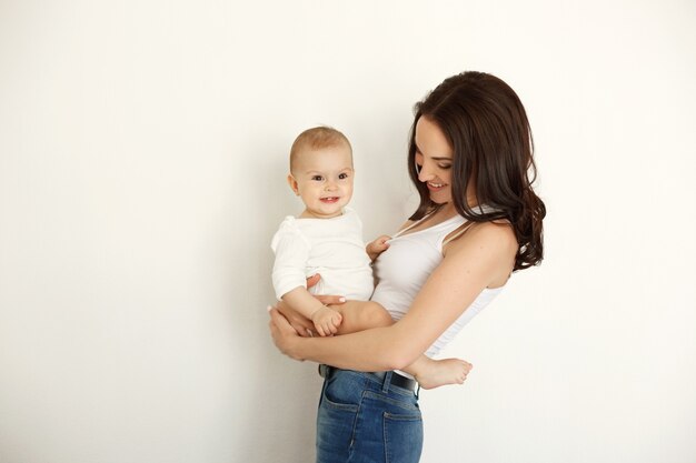 흰 벽 위에 그녀의 아기 딸을 잡고 웃 고 젊은 아름 다운 행복 한 어머니.