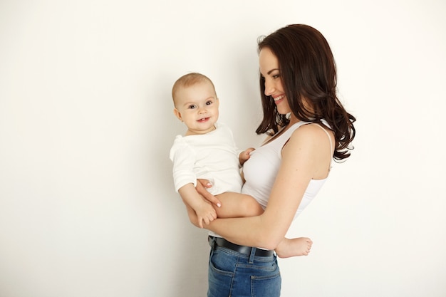 Young beautiful happy mother smiling laughing holding her baby daughter over white wall.